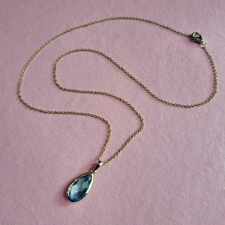 Gold pear drop pendant necklace with blue topaz on a pink background