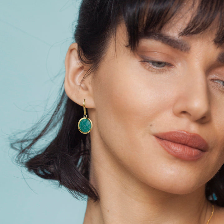 Close-up of a woman wearing a gold amazonite earring with a green gemstone against a light blue background