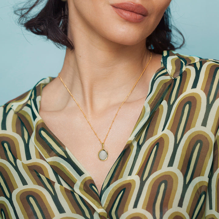 Woman wearing a gold moonstone necklace with a white gemstone close-up of neck and patterned top.