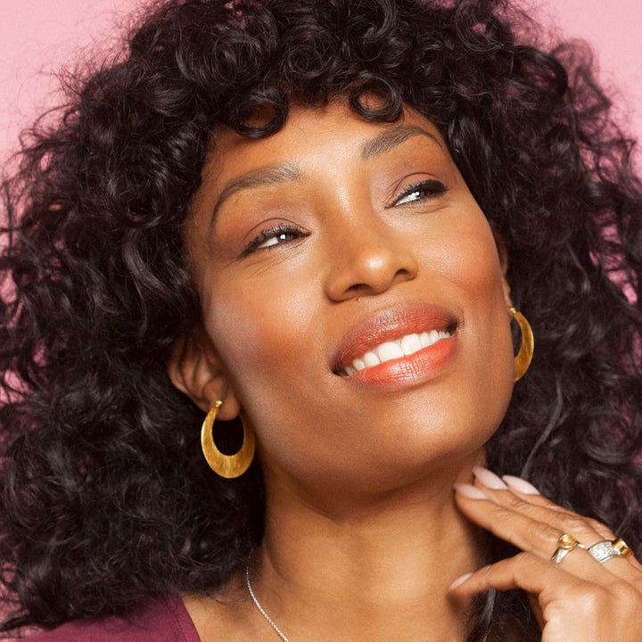 Woman with curly hair wearing gold hoop earrings against a pink background