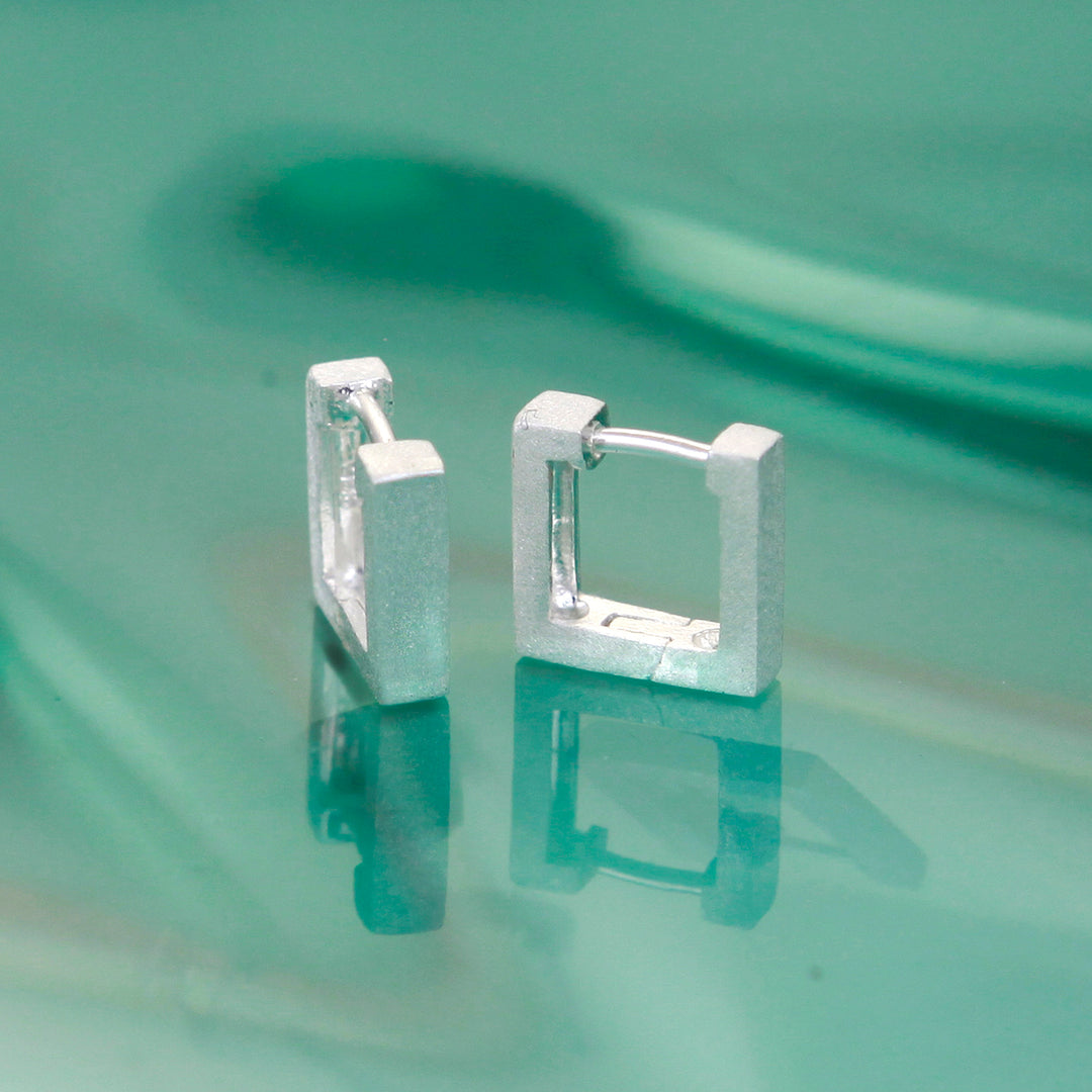 Square silver huggie hoop earrings on a reflective green surface