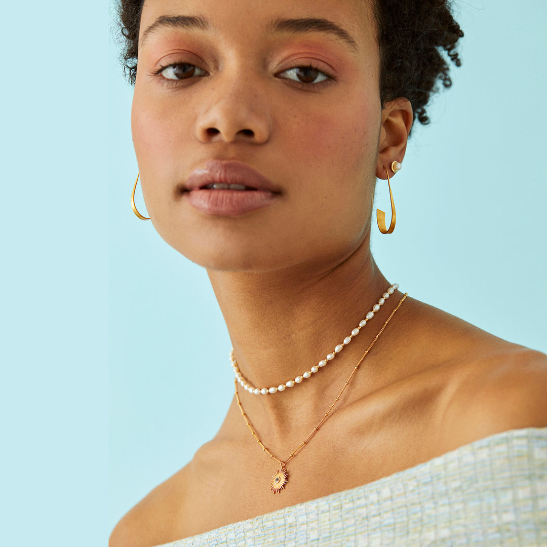 Woman wearing gold threader hoop earrings and pearl necklaces against a light blue background
