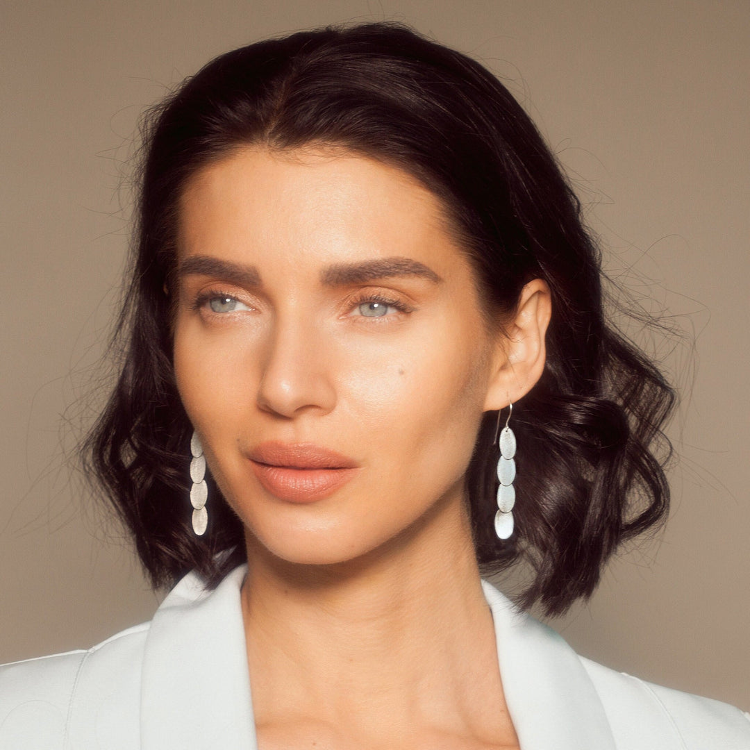 A close-up portrait of a woman with dark wavy hair wearing white oval drop earrings and a white blazer against a warm beige background.