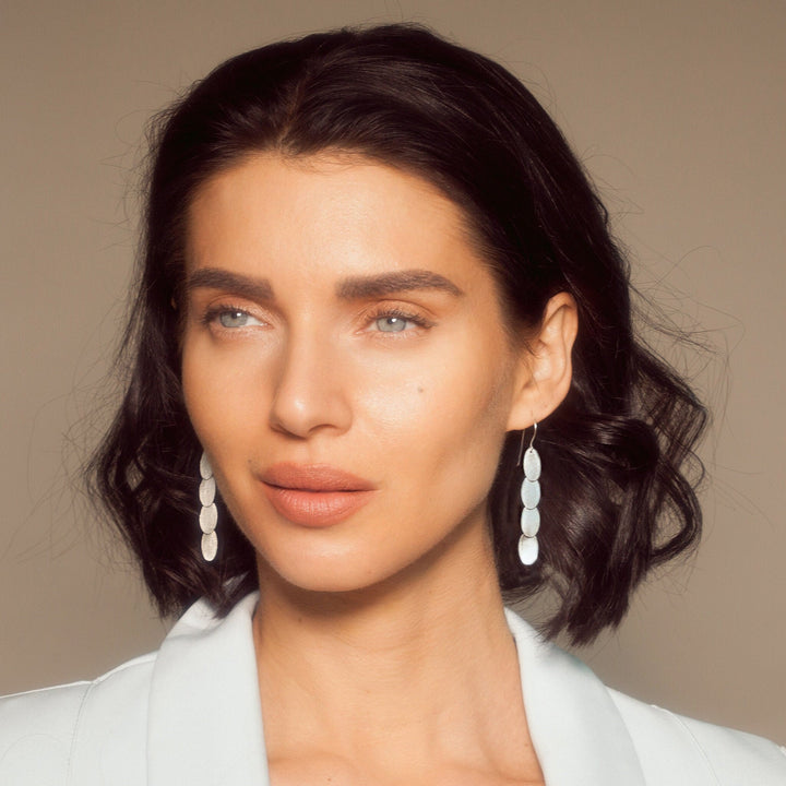 A close-up portrait of a woman with dark wavy hair wearing white oval drop earrings and a white blazer against a warm beige background.