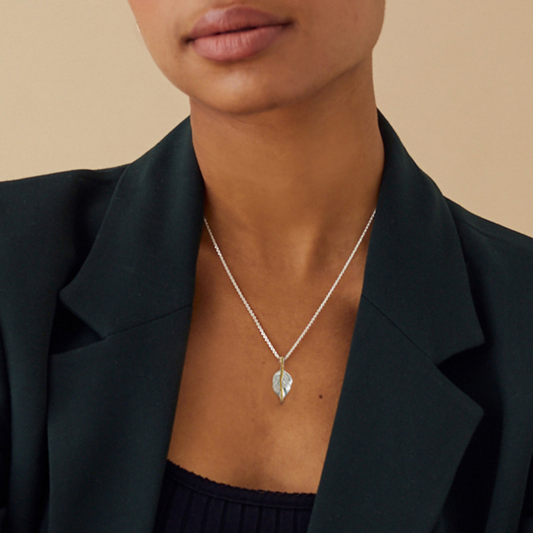 Person wearing a silver and gold necklace with a leaf pendant against a beige background