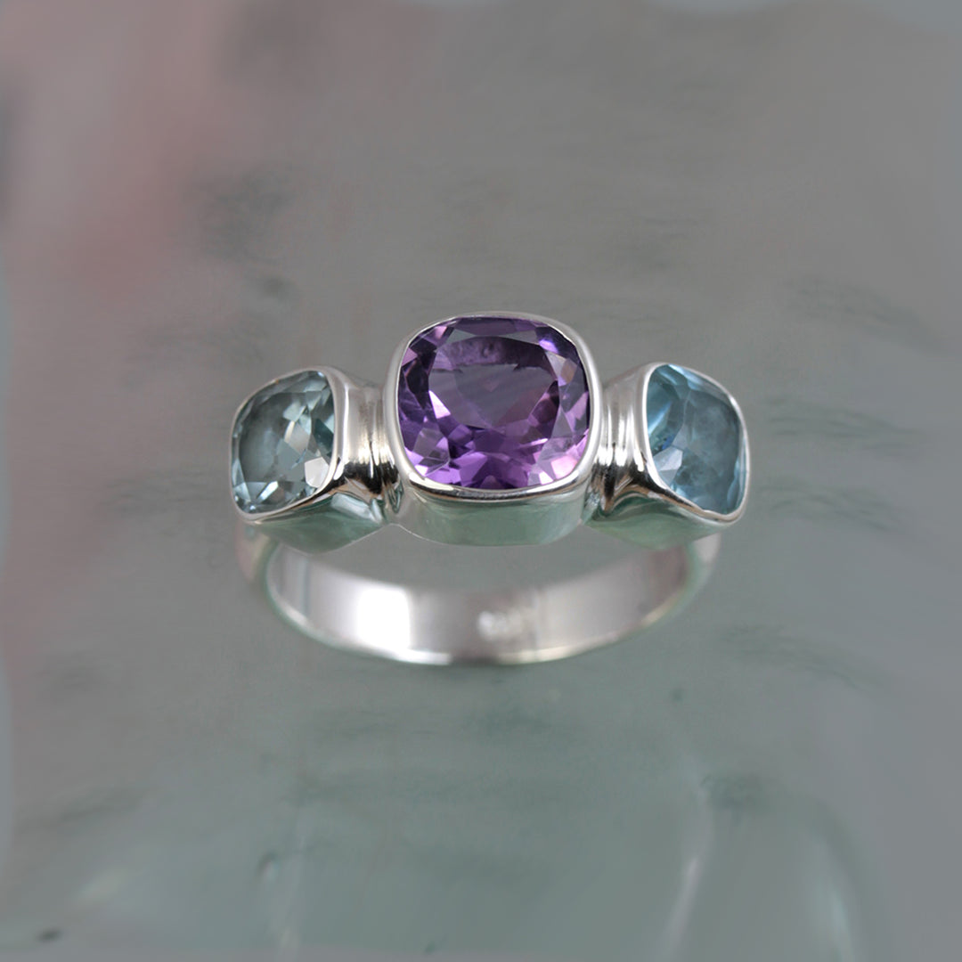 Front view of silver ring with square amethyst and blue topaz gemstones on a glass surface