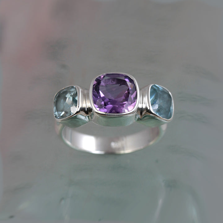 Front view of silver ring with square amethyst and blue topaz gemstones on a glass surface