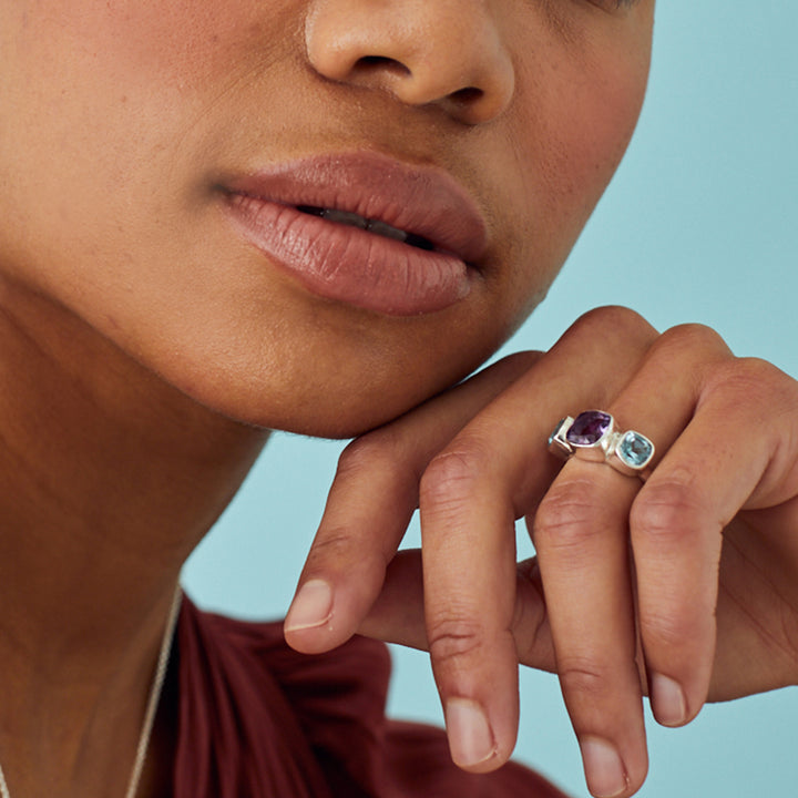 Model wearing a silver ring set with amethyst and blue topaz gemstones on a turquoise background