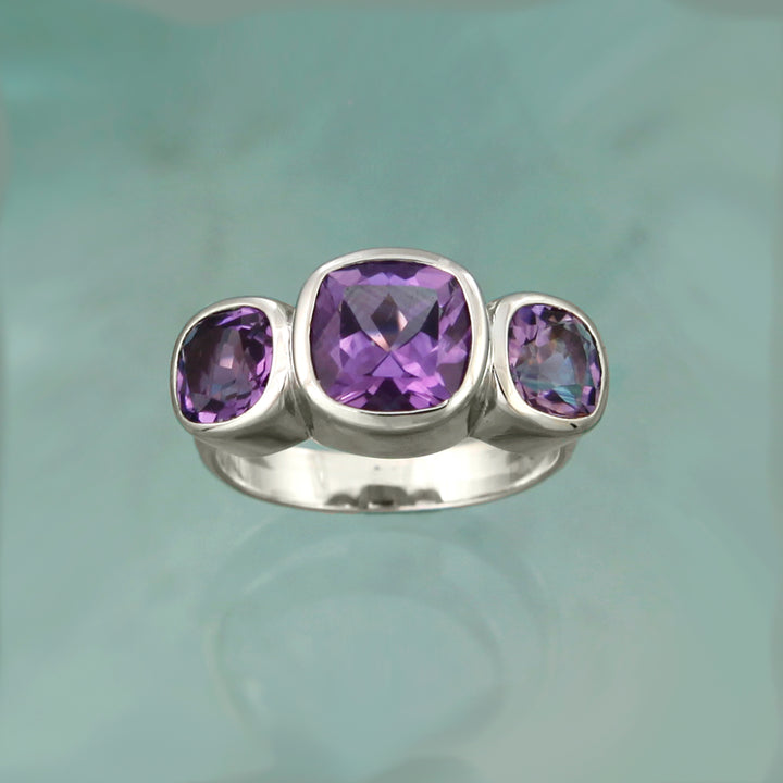front view of a silver ring set with three square purple amethysts on an aqua background