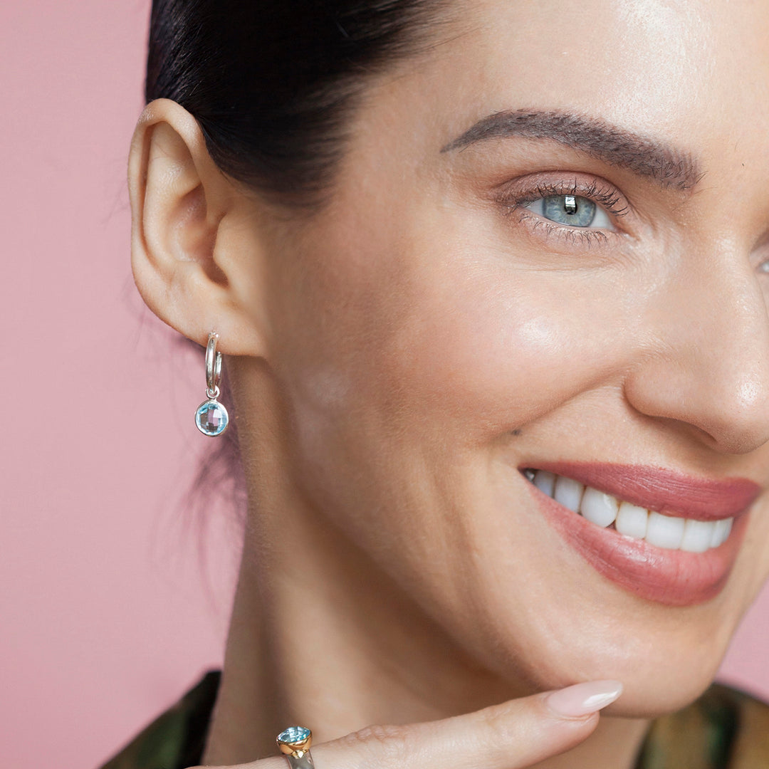 Close-up of model wearing silver huggie hoop earrings with blue topaz drops