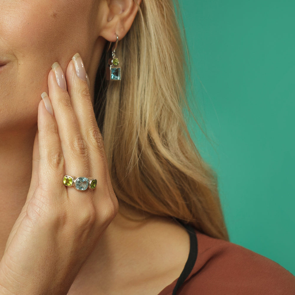 Model wearing silver ring with blue topaz and peridot gems with matching earrings on green background