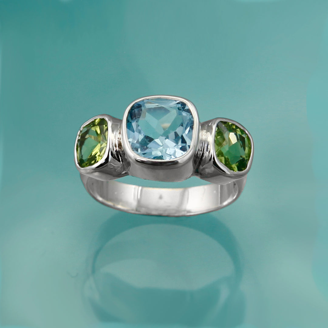Three stone ring in silver with blue and green gemstones on an aqua background