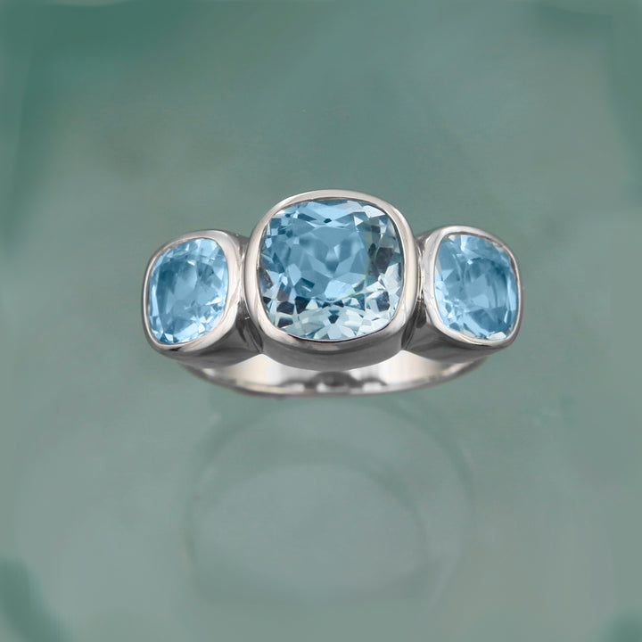 Front  view of a three cushion-cut blue topaz ring on aqua background