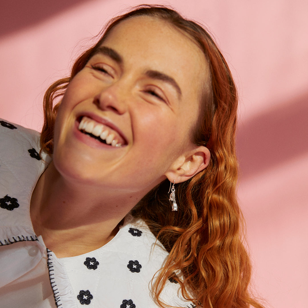 Woman wearing silver flower earrings with red hair smiling against a pink background