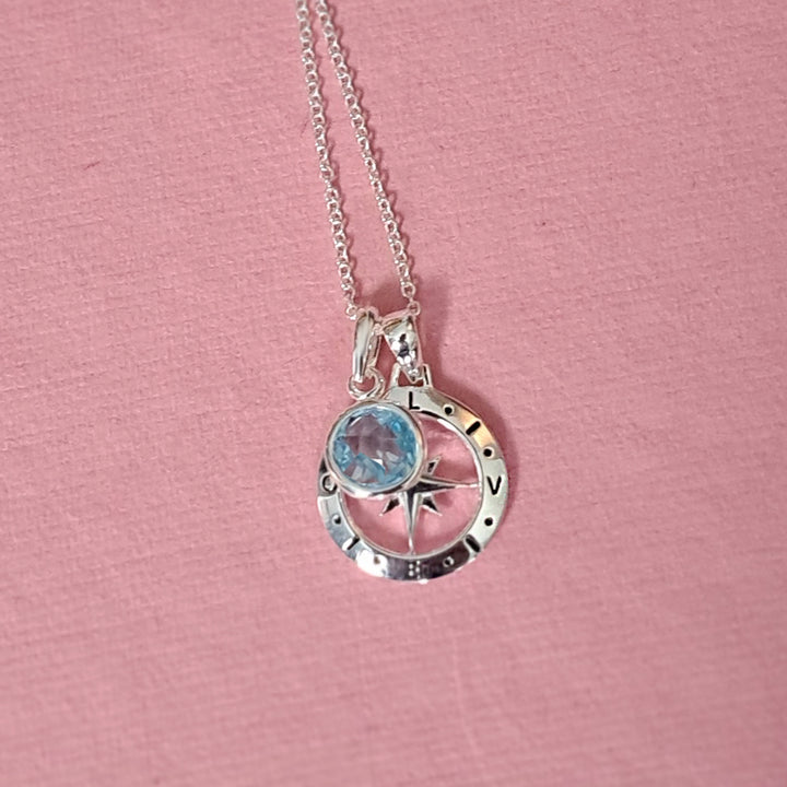 Close-up of silver compass necklace with birthstone charm on pink background