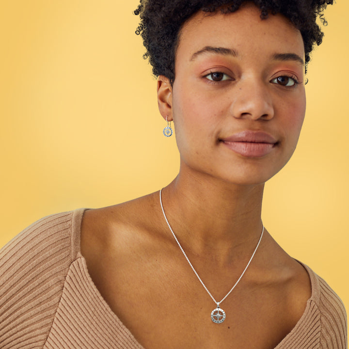 Woman wearing a silver compass earrings and matching necklace with a yellow background