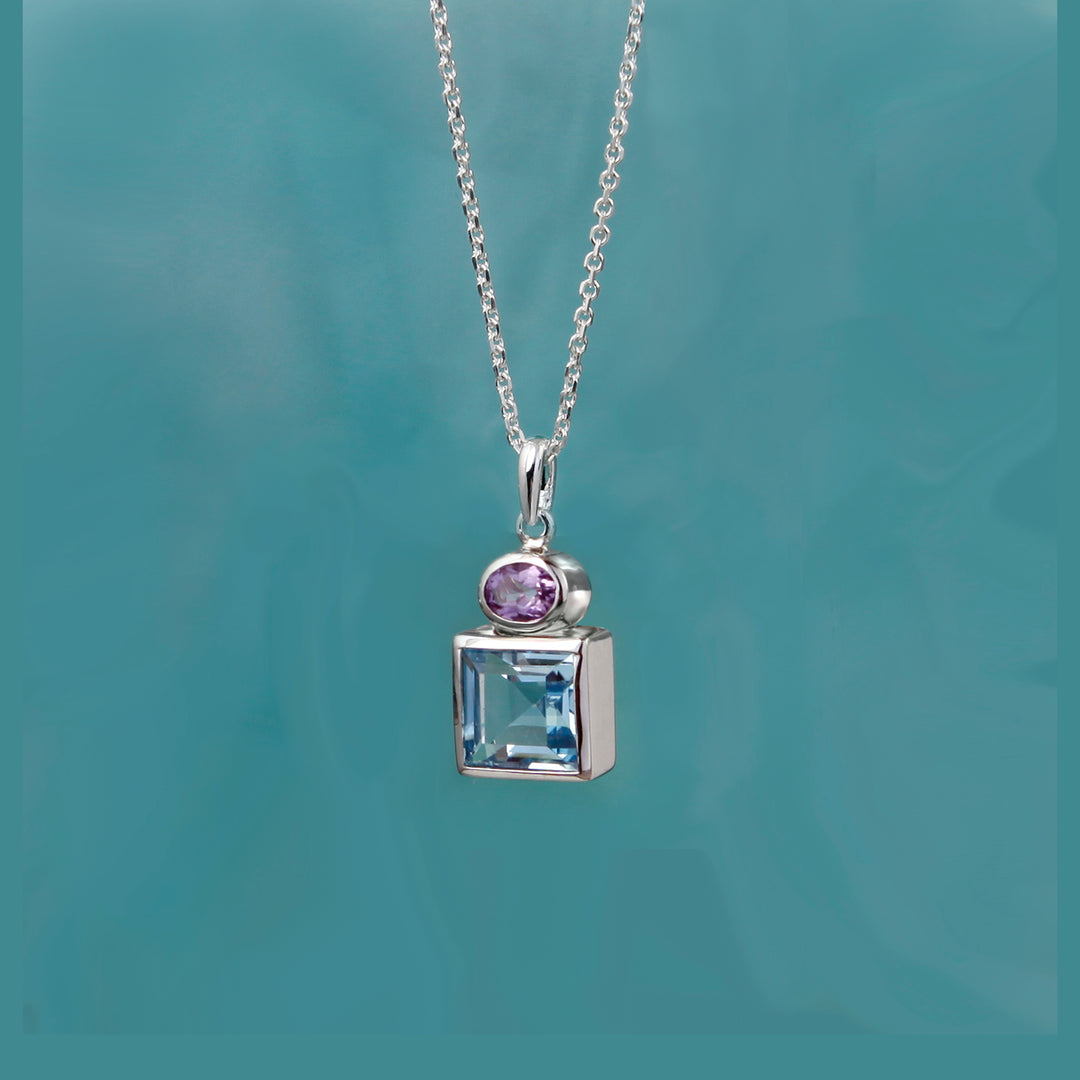 Silver necklace with a pendant featuring a purple amethyst gemstone and square-cut blue topaz on a teal background