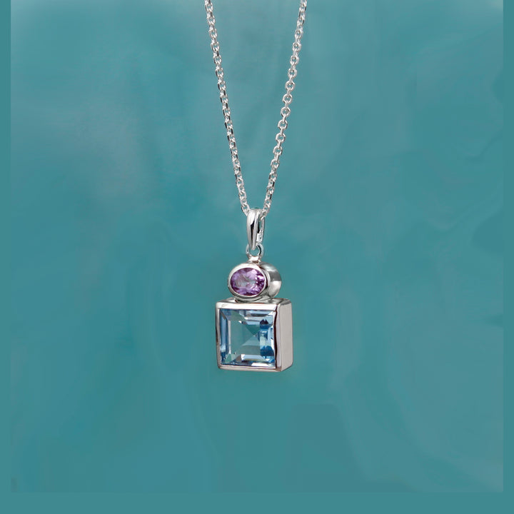 Silver necklace with a pendant featuring a purple amethyst gemstone and square-cut blue topaz on a teal background