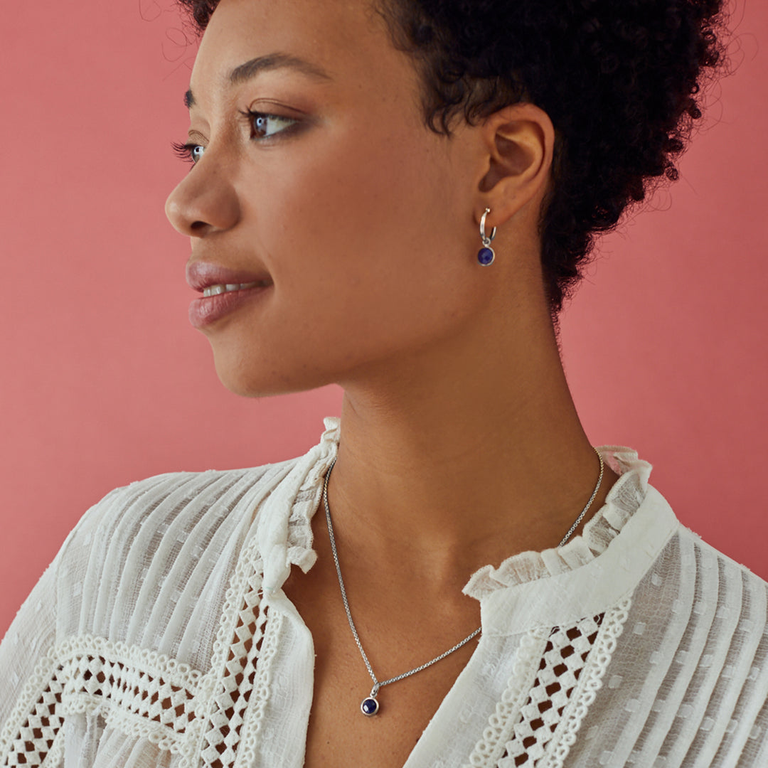 Woman wearing silver huggie earrings with matching pendant set with navy blue gemstones.