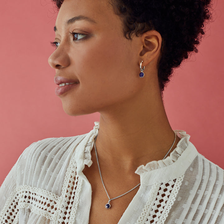 Woman wearing silver huggie earrings with matching pendant set with navy blue gemstones.