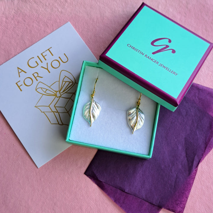 A pair of gold and silver leaf drop earrings displayed in an open Christin Ranger Jewellery box with purple tissue and a gift note.