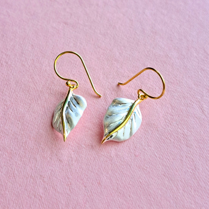 Silver leaf earrings in a matte finish with gold stem and hooks on a pink background.