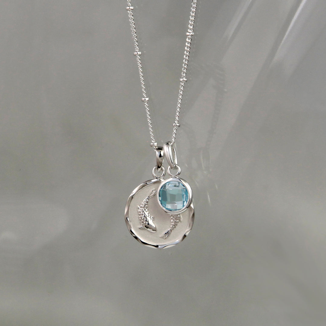 Silver Pisces pendant and blue topaz march birthstone charm on a gray background