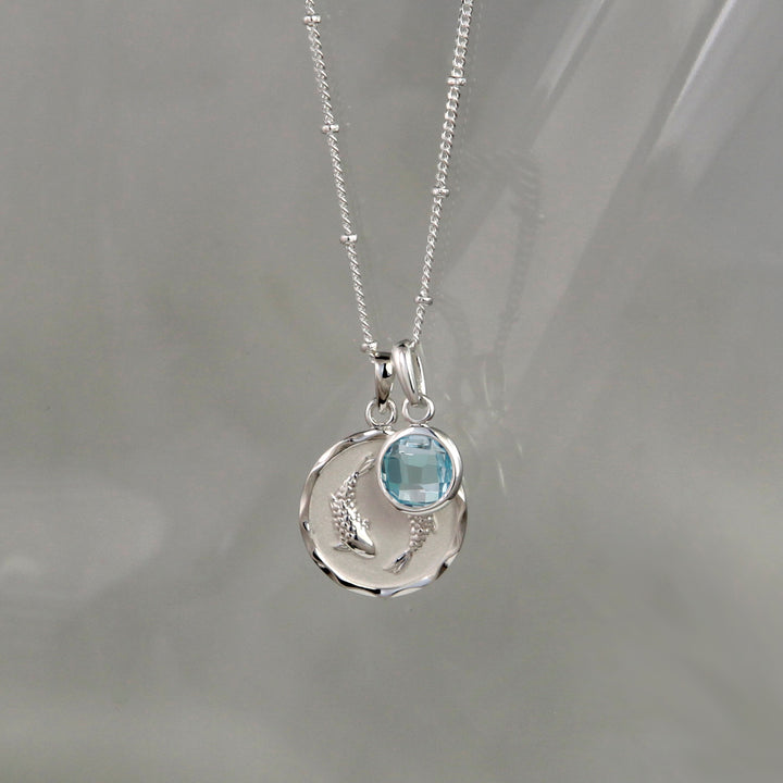 Silver Pisces pendant and blue topaz march birthstone charm on a gray background
