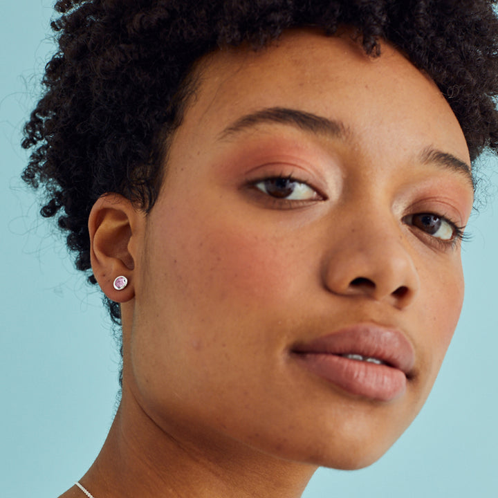 Model wearing minimal silver stud earrings with pale pink gemstones on a blue background.