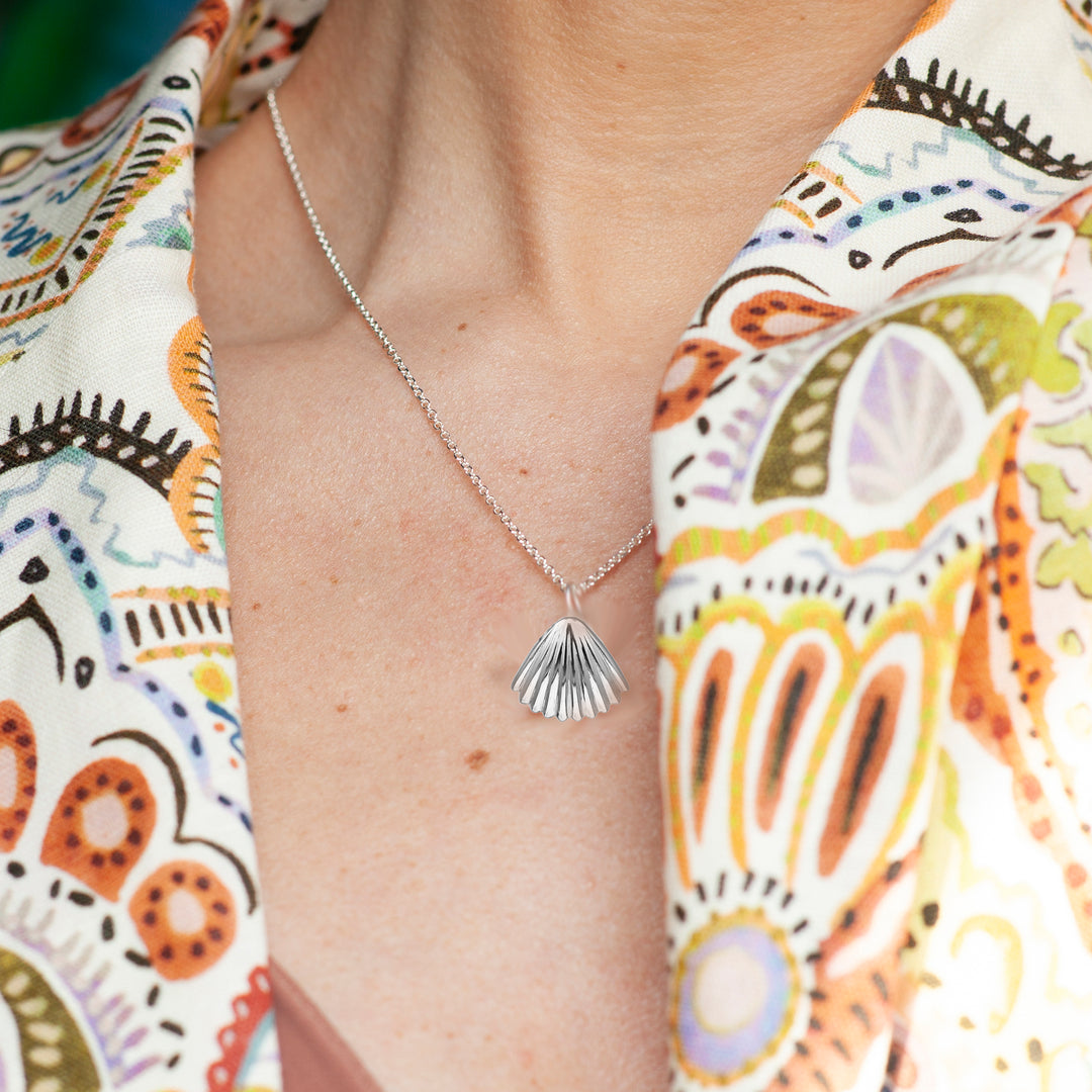 Person wearing a silver necklace with a shell pendant against a colorful patterned fabric background