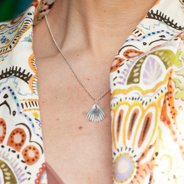 Person wearing a silver necklace with a shell pendant against a colorful patterned fabric background