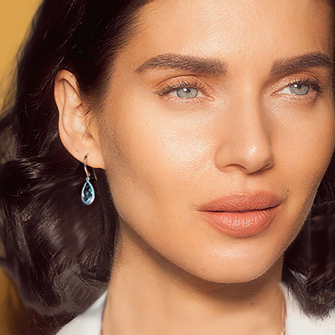 Close-up of woman wearing teardrop shaped blue topaz gemstone earrings on hooks.