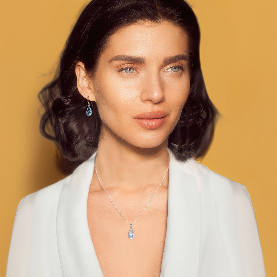 Image of woman wearing silver teardrop earring and necklace set in blue topaz on a yellow background.