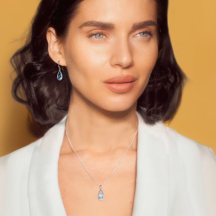 A close-up portrait of a woman wearing a delicate silver necklace with a teardrop-shaped light blue gemstone pendant and a matching drop earring, set against a solid warm yellow background.