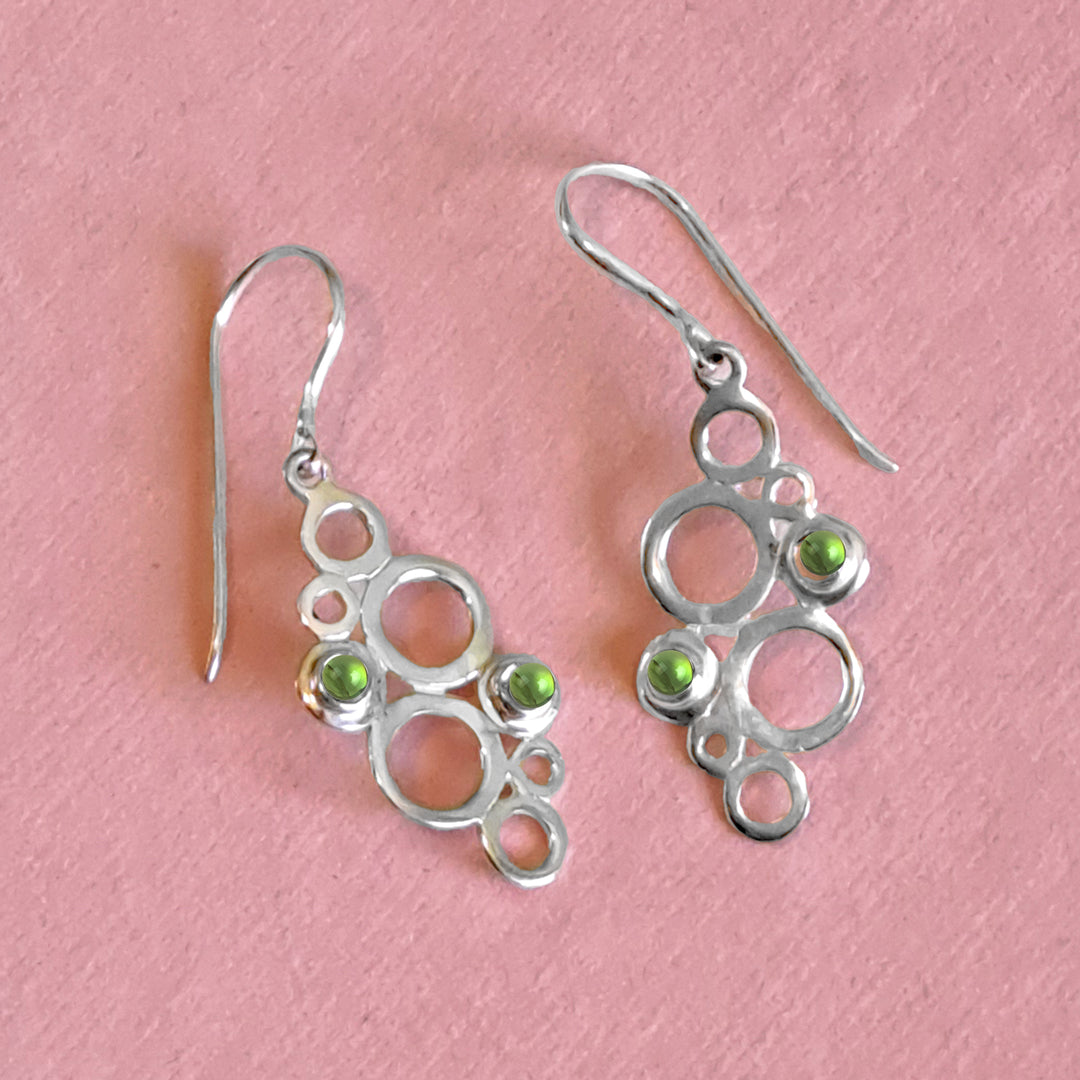 Sterling silver geometric drop earrings with green peridot cabochon gemstones on a pink background