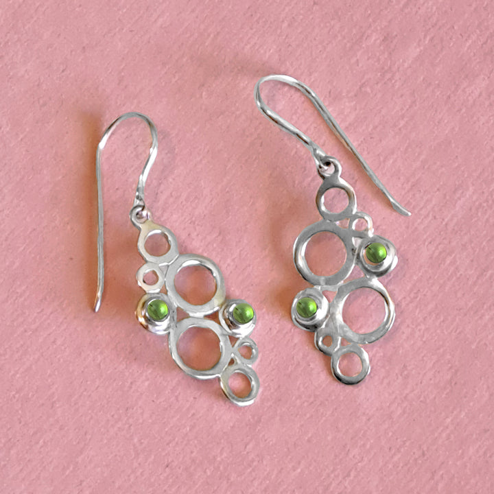 Sterling silver geometric drop earrings with green peridot cabochon gemstones on a pink background