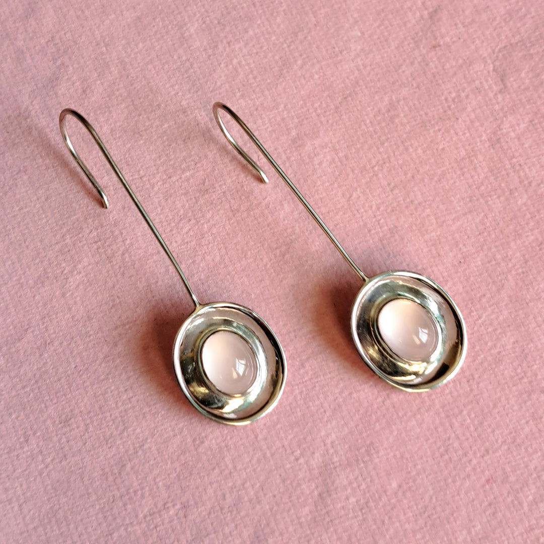 Sterling silver long dish drop earrings with an oval rose quartz cabochon gemstone laying on a pink background
