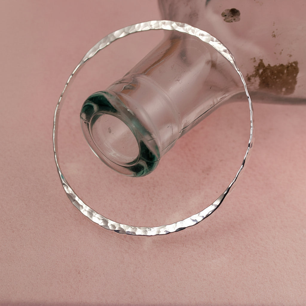 Sterling silver double take bangle with hammered texture with a bottle on a pink background