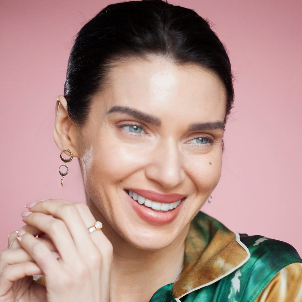 Woman wearing gold and silver dangle earrings and a ring against a pink background