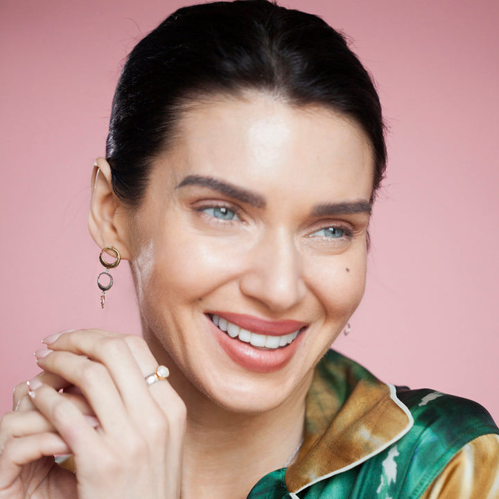 Woman wearing gold and silver dangle earrings and a ring against a pink background