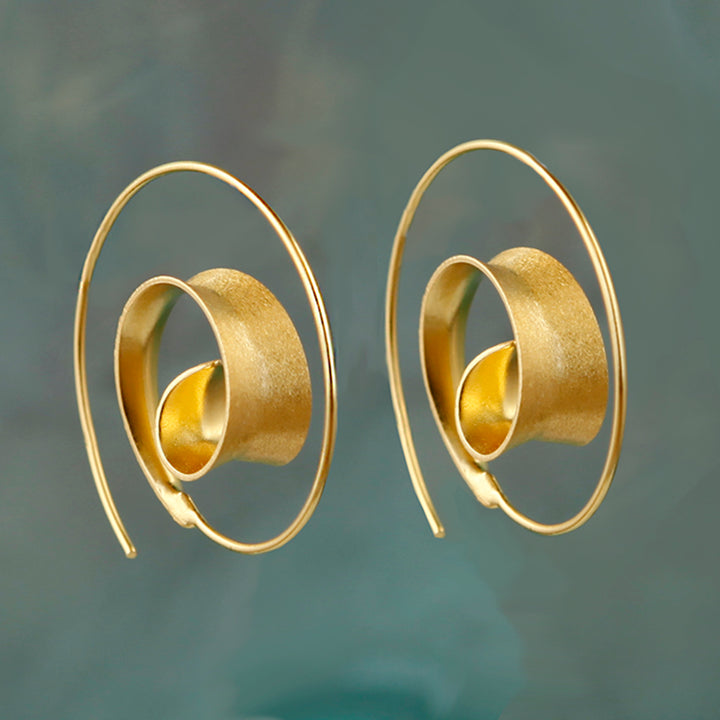 Pair of gold spiral hoop earrings on a reflective surface with a blurred background