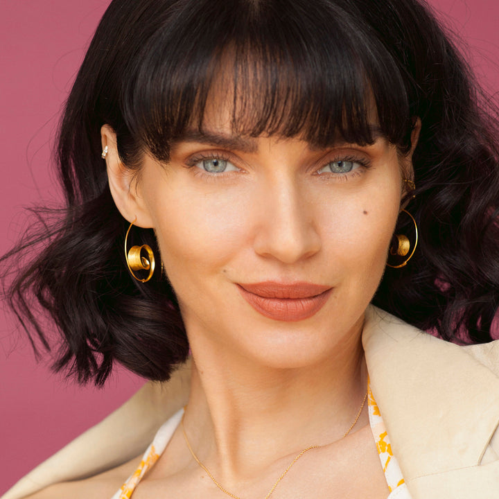 Woman with dark hair and gold threader hoop earrings against a pink background
