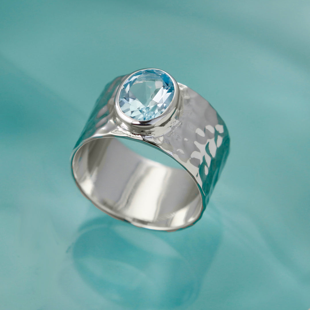 Large blue topaz gemstone set in a wide hammered silver band on an aqua background