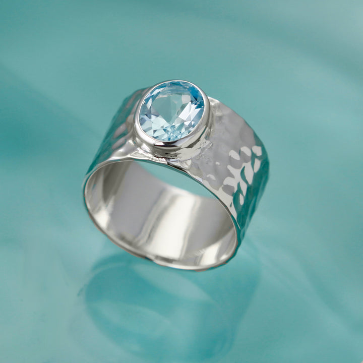 Large blue topaz gemstone set in a wide hammered silver band on an aqua background