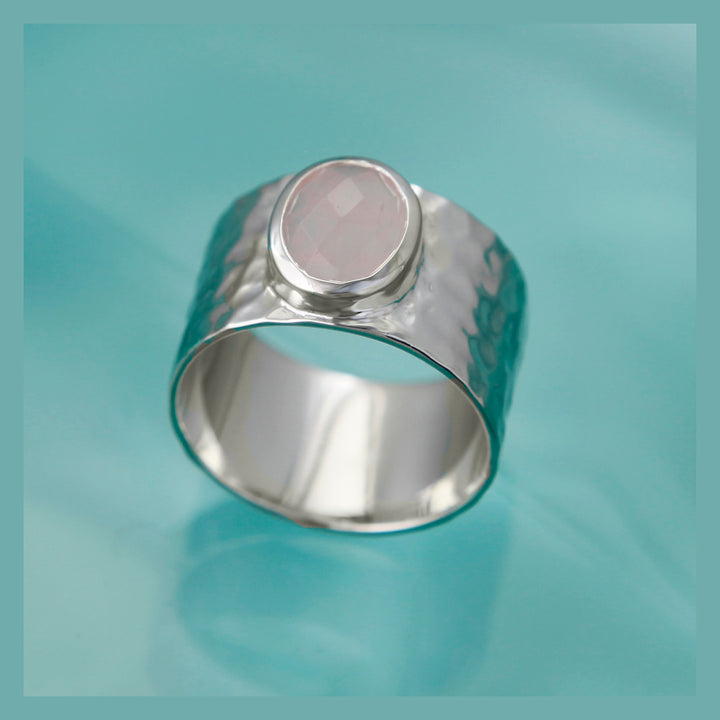 Wide silver ring with a hammered texture set with a rose quartz stone on an aqua background