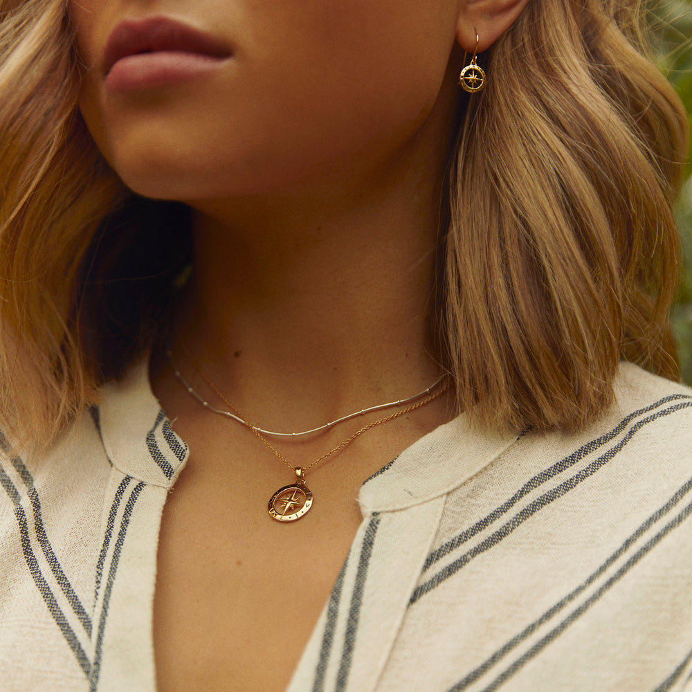 Close-up of model wearing a gold compass necklace and chain with matching earrings.